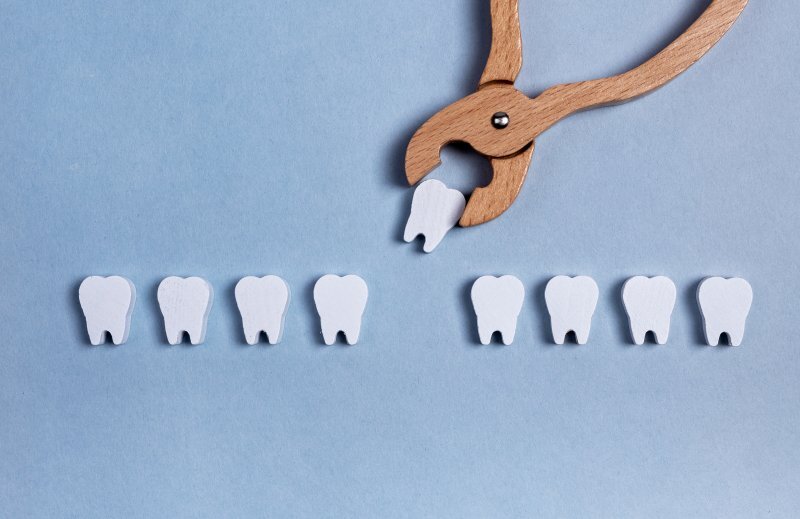 Wooden dental pliers extracting a tooth from a row of eight white tooth models on blue background.