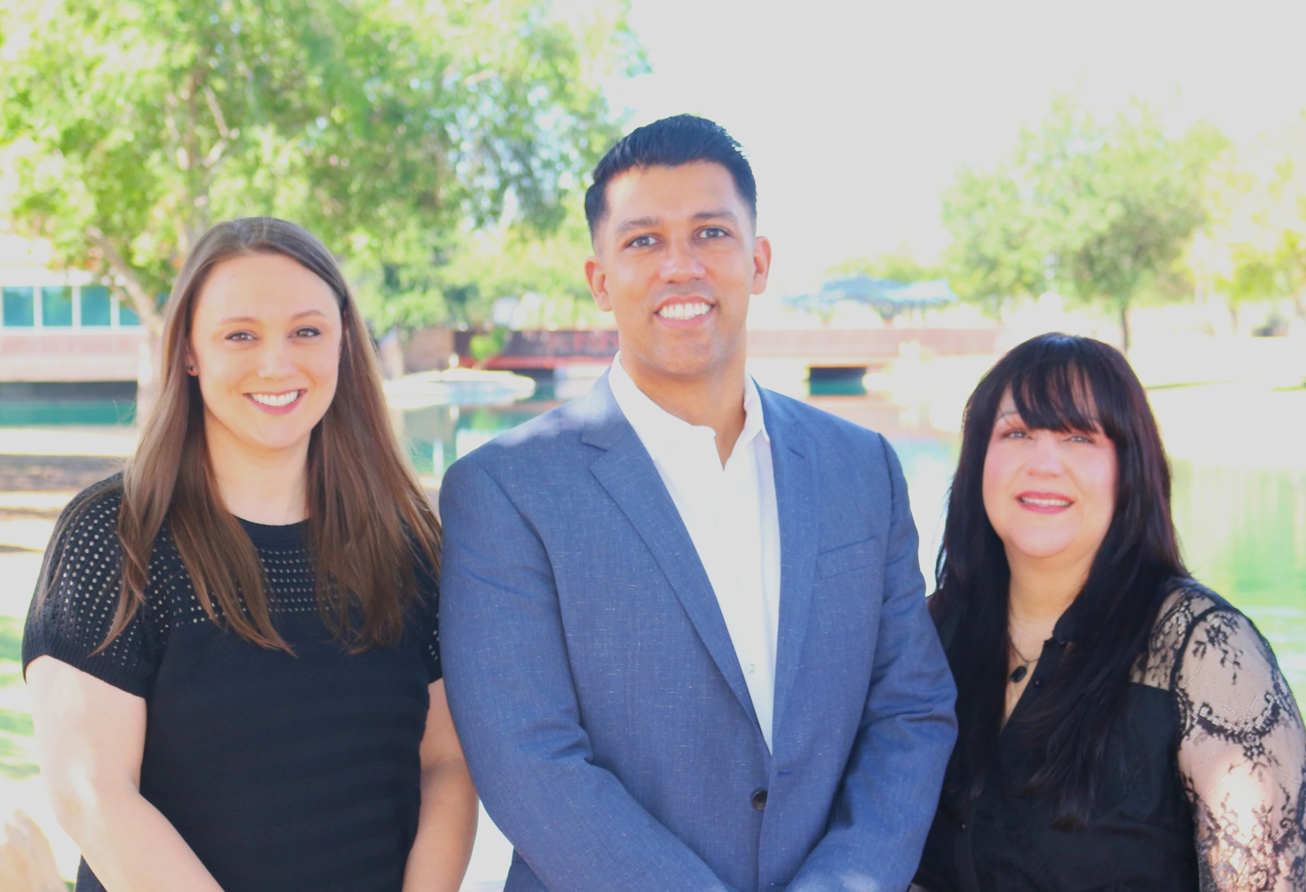 Three smiling team members standing close together outdoors, a man in a blue blazer between two women in black, with trees and a pond behind them